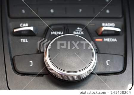 Close-up view of modern car infotainment system control panel, displaying large central rotary dial with textured metal edge, surrounded by buttons for various vehicle functions. Close-up view of modern car infotainment system control panel, displaying large central rotary dial with textured metal edge, surrounded by buttons for various vehicle functions. 141438264