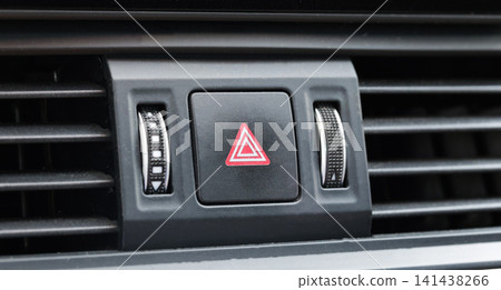 Vehicle dashboard close-up showing hazard warning light button with red triangle symbol, surrounded by car interior air conditioning vents and rotary controls for safety feature Vehicle dashboard close-up showing hazard warning light button with red triangle symbol, surrounded by car interior air conditioning vents and rotary controls for safety feature 141438266