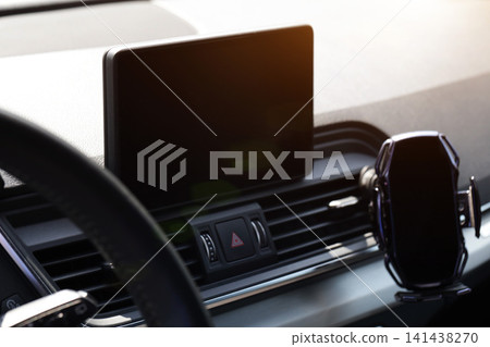 Modern car dashboard detail showing black infotainment screen, hazard warning button, and smartphone mount with air conditioning vents, sunlight flare creating reflection. 141438270