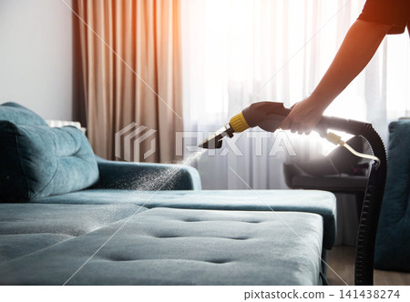 Cleaning blue upholstered sofa with wet vacuum cleaner in living room, hand spraying cleaning solution for deep fabric sanitation and stain removal Cleaning blue upholstered sofa with wet vacuum cleaner in living room, hand spraying cleaning solution for deep fabric sanitation and stain removal 141438274