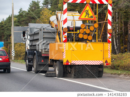 Road sweeper truck with warning sign and hazard stripes cleaning edge of asphalt road with forest background Road sweeper truck with warning sign and hazard stripes cleaning edge of asphalt road with forest background 141438283