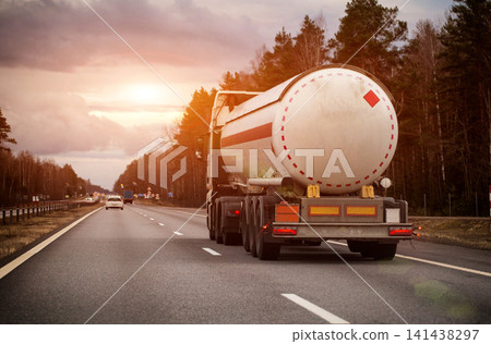 White tanker truck transporting flammable liquid on multi-lane highway surrounded by pine forest and bare trees under overcast sky, evening sunset. Copy space for text White tanker truck transporting flammable liquid on multi-lane highway surrounded by pine forest and bare trees under overcast sky, evening sunset. Copy space for text 141438297