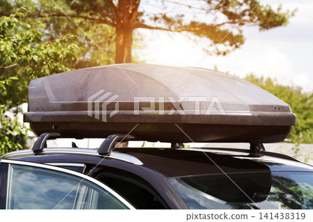 Dark color automobile with roof cargo box mounted on roof racks, parked outdoors under bright sunlight, surrounded by green trees and natural environment, clear day. Dark color automobile with roof cargo box mounted on roof racks, parked outdoors under bright sunlight, surrounded by green trees and natural environment, clear day. 141438319