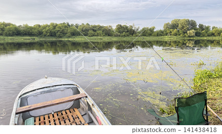 A fishing spot on a quiet river: a boat near the shore, a fishing rod, and a folding chair in front of a green forest. A fishing spot on a quiet river: a boat near the shore, a fishing rod, and a folding chair in front of a green forest. 141438418