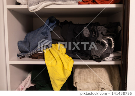 Open wardrobe shelves with casually stacked clothes, bags and a bright yellow garment, showing cluttered everyday storage and realistic home organization in a domestic interior Open wardrobe shelves with casually stacked clothes, bags and a bright yellow garment, showing cluttered everyday storage and realistic home organization in a domestic interior 141438756