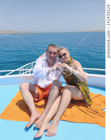 A happy couple on a sunlit yacht frames a heart with their hands against the backdrop of a serene blue sea, radiating love, connection, and the joy of shared moments during a dreamy coastal getaway. A happy couple on a sunlit yacht frames a heart with their hands against the backdrop of a serene blue sea, radiating love, connection, and the joy of shared moments during a dreamy coastal getaway. 141439128