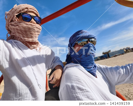 Father and son share an exhilarating ride dune buggy across golden desert sandswith both wearing traditional headscarves and sunglasses, capturing joy thrilling family adventure under bright blue sky Father and son share an exhilarating ride dune buggy across golden desert sandswith both wearing traditional headscarves and sunglasses, capturing joy thrilling family adventure under bright blue sky 141439159