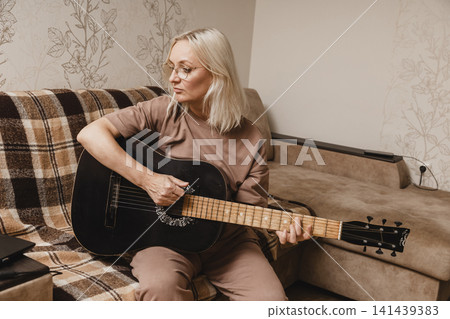Mature woman playing acoustic guitar in her living room Mature woman playing acoustic guitar in her living room 141439383