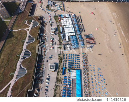 Beautiful high altitude aerial panoramic perspective of beach resort clubs with blue sunbeds green roof paths and solar panels along a seaside promenade under bright sunlight Beautiful high altitude aerial panoramic perspective of beach resort clubs with blue sunbeds green roof paths and solar panels along a seaside promenade under bright sunlight 141439826