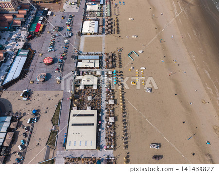 Beautiful high altitude aerial panoramic perspective of beach resort clubs with straw parasols promenade boulevard vintage cars and sandy ocean coast under bright sunlight Beautiful high altitude aerial panoramic perspective of beach resort clubs with straw parasols promenade boulevard vintage cars and sandy ocean coast under bright sunlight 141439827