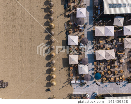 Beautiful top down high altitude aerial panoramic perspective of beach club terrace with straw parasols white umbrellas and wooden outdoor furniture under bright sunlight Beautiful top down high altitude aerial panoramic perspective of beach club terrace with straw parasols white umbrellas and wooden outdoor furniture under bright sunlight 141439828