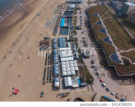 Beautiful high altitude aerial panoramic perspective of beach resort clubs with blue sunbeds green roof solar panels and coastal promenade along the sandy shoreline under bright sunlight Beautiful high altitude aerial panoramic perspective of beach resort clubs with blue sunbeds green roof solar panels and coastal promenade along the sandy shoreline under bright sunlight 141439829
