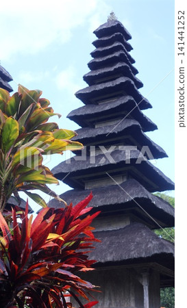 Tiered Balinese temple roof rising above tropical foliage under bright sky, with stacked thatch silhouette, colorful croton leaves, island heritage, calm outdoor scene Tiered Balinese temple roof rising above tropical foliage under bright sky, with stacked thatch silhouette, colorful croton leaves, island heritage, calm outdoor scene 141441252