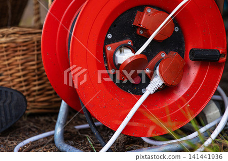 Closeup of a red power cable reel with extension cords plugged in, set outdoors on the ground. Practical solution for electricity in nature remote areas Closeup of a red power cable reel with extension cords plugged in, set outdoors on the ground. Practical solution for electricity in nature remote areas 141441936