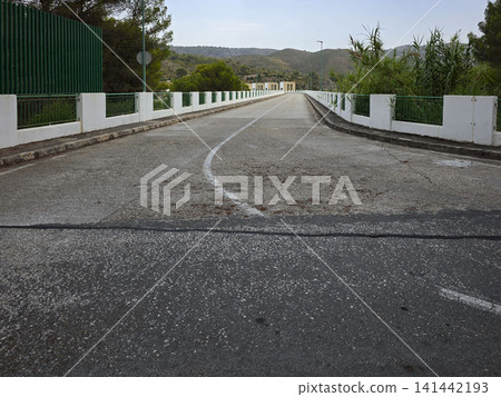 The concrete road runs along the top of the Beniarres Reservoir dam wall. White safety barriers line the edges under a cloudy sky. Green hills are visible in the background. The concrete road runs along the top of the Beniarres Reservoir dam wall. White safety barriers line the edges under a cloudy sky. Green hills are visible in the background. 141442193