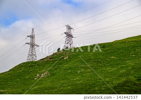 Electricity transmission towers on green hill against overcast sky Electricity transmission towers on green hill against overcast sky 141442537