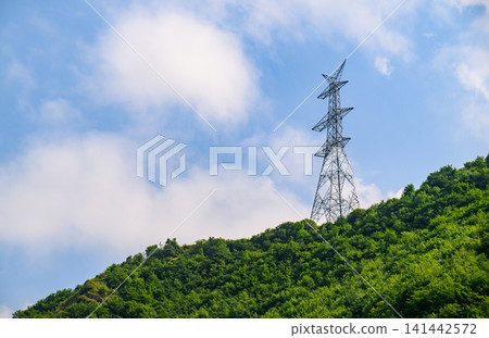 Power line tower rises above forested mountain peak beneath partly cloudy sky Power line tower rises above forested mountain peak beneath partly cloudy sky 141442572