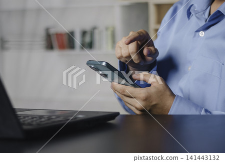 Businessman using smartphone while working on a laptop, representing mobile technology, digital communication, online productivity and remote workflow in a modern office environment. Life style Businessman using smartphone while working on a laptop, representing mobile technology, digital communication, online productivity and remote workflow in a modern office environment. Life style 141443132