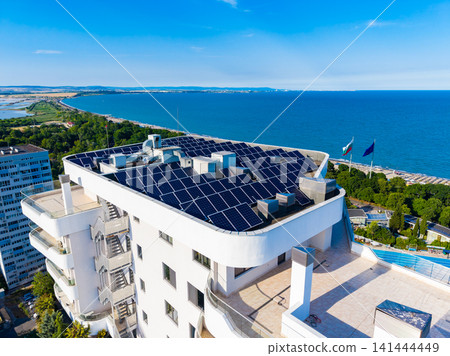 Solar panels on a rooftop overlooking the sea with a clear sky and a coastal view in the background during daylight hours Solar panels on a rooftop overlooking the sea with a clear sky and a coastal view in the background during daylight hours 141444449