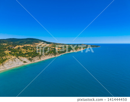 High-angle drone view of rugged coastal cliffline meeting clear blue sea under bright sky, with sunlit scrubby slopes, sandy coves, and long uninterrupted shoreline High-angle drone view of rugged coastal cliffline meeting clear blue sea under bright sky, with sunlit scrubby slopes, sandy coves, and long uninterrupted shoreline 141444450