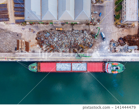 Cargo ship unloading materials at a dock near a recycling site with piles of waste and nearby buildings in an industrial area Cargo ship unloading materials at a dock near a recycling site with piles of waste and nearby buildings in an industrial area 141444479