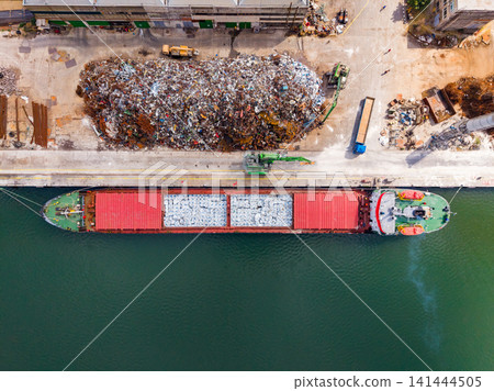 Cargo ship docks at the port to unload materials next to a large pile of scrap metal and industrial equipment 141444505