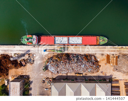 Cargo ship docks at port to unload materials near recycling area along the water during daytime Cargo ship docks at port to unload materials near recycling area along the water during daytime 141444507