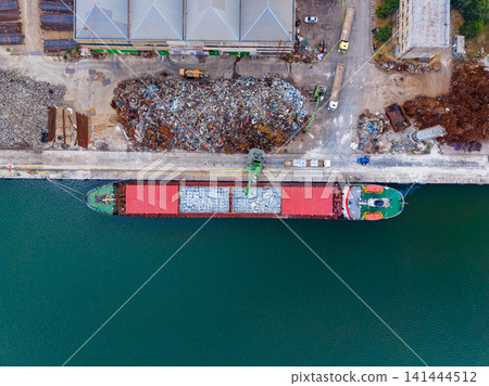 Commercial shipping vessel docked at an industrial port with loading equipment and scrap material nearby 141444512