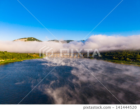 Fog covers the river as the sun rises over the mountains near the bridge creating a scene of nature's beauty in early morning light Fog covers the river as the sun rises over the mountains near the bridge creating a scene of nature's beauty in early morning light 141444520