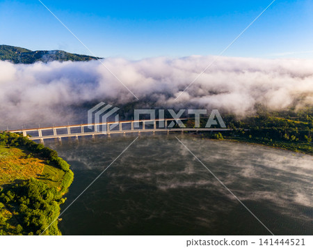 Fog covers a river and bridge in a valley surrounded by mountains in the morning light Fog covers a river and bridge in a valley surrounded by mountains in the morning light 141444521