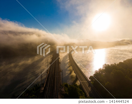 Fog covers the river and train tracks at sunrise in a rural area near the mountains and the water with a clear sky above Fog covers the river and train tracks at sunrise in a rural area near the mountains and the water with a clear sky above 141444524