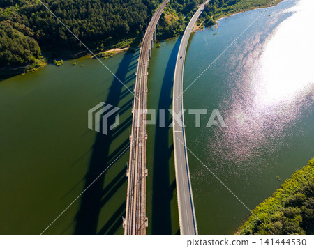 Bridge crosses over a river while a road runs parallel along the water in a green landscape on a clear day Bridge crosses over a river while a road runs parallel along the water in a green landscape on a clear day 141444530
