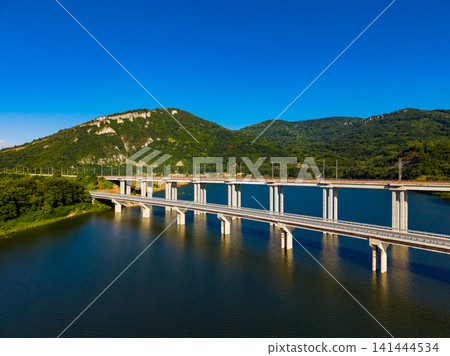 Bridge crosses a large body of water with green mountains in the background under a clear blue sky 141444534