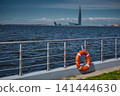 An orange-colored lifebuoy hangs on the embankment fence in sunny weather, finland gulf, with the sea and a skyscraper in the background. 141444630