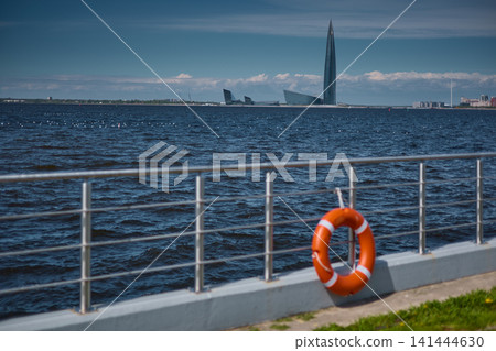 An orange-colored lifebuoy hangs on the embankment fence in sunny weather, finland gulf, with the sea and a skyscraper in the background. 141444630