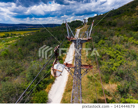 View of a tall transmission tower near a dirt path surrounded by bushes and trees on a sunny day with clouds in the sky View of a tall transmission tower near a dirt path surrounded by bushes and trees on a sunny day with clouds in the sky 141444679