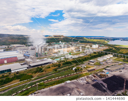 View of a large industrial complex with tall structures and green areas near a river under a cloudy sky in a rural setting View of a large industrial complex with tall structures and green areas near a river under a cloudy sky in a rural setting 141444690