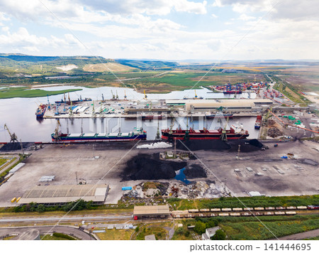 Shipping activities occur at a busy port near a river with multiple vessels loading and unloading goods during daylight hours Shipping activities occur at a busy port near a river with multiple vessels loading and unloading goods during daylight hours 141444695