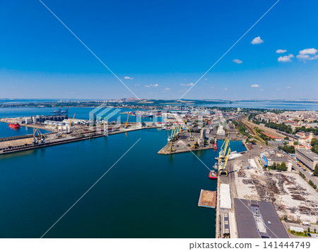 Harbor area with cranes and ships near a city under a clear blue sky during the day Harbor area with cranes and ships near a city under a clear blue sky during the day 141444749