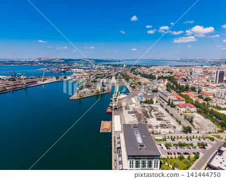 Overlooking a busy port area with cranes, ships, and a city skyline under a bright blue sky during daylight Overlooking a busy port area with cranes, ships, and a city skyline under a bright blue sky during daylight 141444750