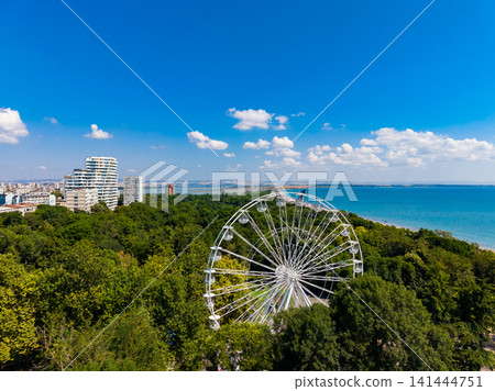 View of a ferris wheel near the sea with buildings and trees in the background on a sunny day View of a ferris wheel near the sea with buildings and trees in the background on a sunny day 141444751