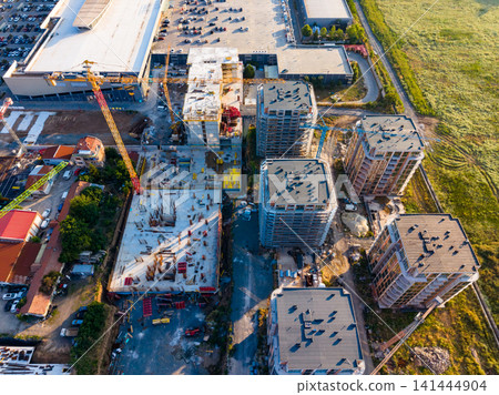Construction site shows multiple buildings being developed near a parking lot with cranes and heavy machinery present in the area 141444904