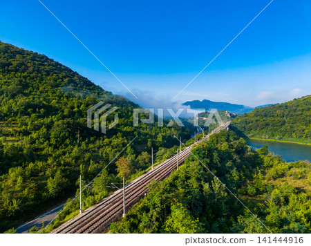 Train tracks run through a green landscape with hills and a river under a clear blue sky in a rural area Train tracks run through a green landscape with hills and a river under a clear blue sky in a rural area 141444916