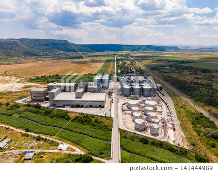 View of a large industrial facility with silos and tanks located near a green area under a cloudy sky in daytime View of a large industrial facility with silos and tanks located near a green area under a cloudy sky in daytime 141444989