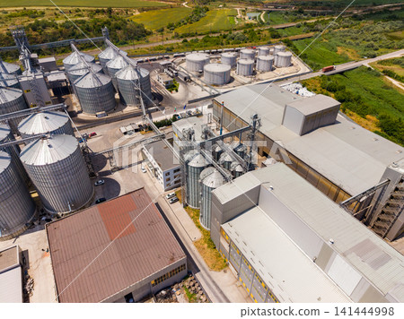 View of a large industrial facility with silos and storage tanks during midday View of a large industrial facility with silos and storage tanks during midday 141444998