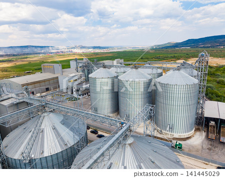 Large storage silos and industrial buildings in a rural area with green fields and cloudy sky Large storage silos and industrial buildings in a rural area with green fields and cloudy sky 141445029