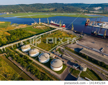 Industrial area with storage tanks and cargo ships along a river during daytime in an expansive landscape Industrial area with storage tanks and cargo ships along a river during daytime in an expansive landscape 141445284