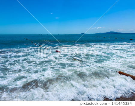 Waves crash on the shore near a fishing area with floating logs and a view of distant islands in bright daylight Waves crash on the shore near a fishing area with floating logs and a view of distant islands in bright daylight 141445324