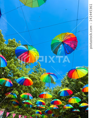 Colorful umbrellas hang over a street in a bright blue sky during a sunny day while people walk underneath them enjoying the festive decoration Colorful umbrellas hang over a street in a bright blue sky during a sunny day while people walk underneath them enjoying the festive decoration 141445361