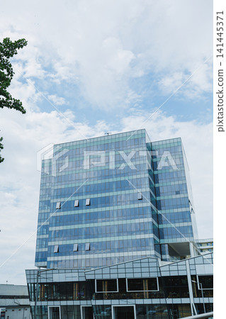 Tall glass building under cloudy sky in urban location showing modern architecture with multiple reflections and clear lines of design Tall glass building under cloudy sky in urban location showing modern architecture with multiple reflections and clear lines of design 141445371